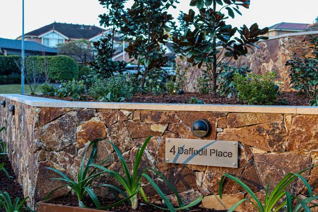 Exterior view of a landscaped stone masonry boundary wall and raised garden bed, featuring a mounted address plaque reading “4 Daffodil Place,” with residential houses and hedges in the background.