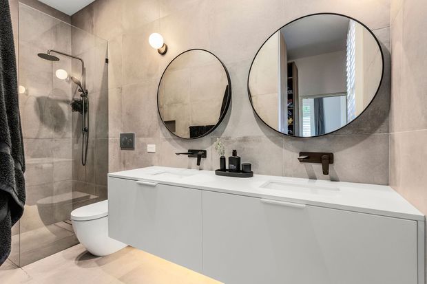 Contemporary bathroom with a long floating white vanity, two integrated basins, matte black wall-mounted tapware, twin round mirrors, and a walk-in glass shower. Large-format warm grey stone-look wall and floor tiles create a refined, neutral finish, with concealed under-vanity lighting.
