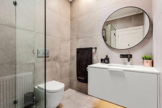 A contemporary bathroom with large beige-grey stone-look wall and floor tiles, a frameless glass shower enclosure, wall-hung white toilet, floating white vanity with integrated basin, black wall-mounted tapware, round black-framed mirror, and dark towel accents. The room is compact, clean, and softly lit.