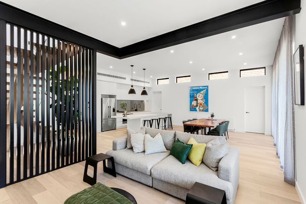 Bright open-plan living space with a large light-grey sectional sofa in the foreground, black side tables, and a partially visible green upholstered ottoman. A black vertical timber-look screen separates the living area from the kitchen. The kitchen has white cabinetry, a pale benchtop, stainless-steel appliances and pendant lights, while a timber dining table sits beyond. Pale timber flooring, white walls, black ceiling beams and high clerestory windows create a clean contemporary interior.