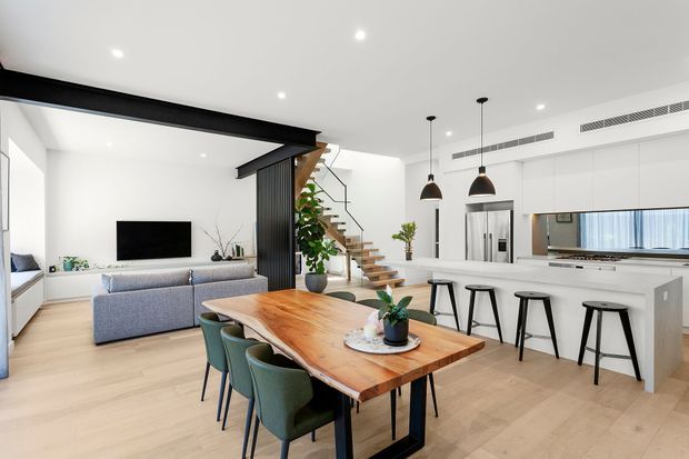 Bright open-plan contemporary living, dining and kitchen area with pale timber flooring, a large natural timber dining table, upholstered green dining chairs, a grey sofa, white cabinetry, a long kitchen island with black stools, pendant lights, and a floating timber stair beside a dark vertical screen. Recessed ceiling lighting and extensive white surfaces create an airy, clean interior.