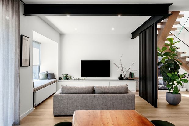 Bright contemporary living room with a grey modular sofa facing a wall-mounted television and low white built-in media cabinetry. A timber coffee table, pale timber flooring, white walls, sheer curtains, black steel overhead beam and slatted room divider create a clean, modern interior. A timber stair with black balustrade and indoor greenery are visible to the right, while a built-in window seat appears at left.