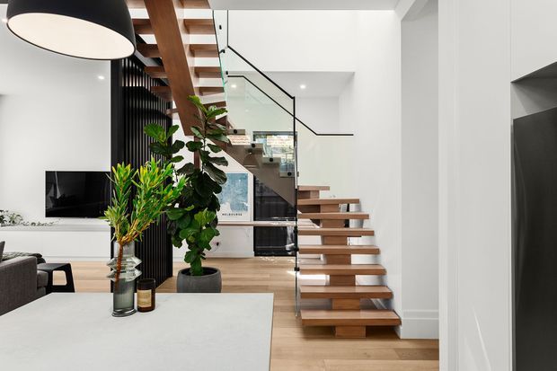 Contemporary open-plan interior featuring a floating timber staircase with clear glass balustrade and slim black handrail. White walls and cabinetry, light timber flooring, black vertical screening, recessed lighting, and a partially visible living area create a clean, bright modern setting. A large island or counter occupies the foreground, with indoor plants and a television visible beyond.