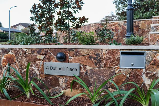 Close-up exterior view of a landscaped residential boundary wall constructed from irregular reddish-brown stone, featuring a metal address plaque, recessed mailbox, black lamp fixture, garden planting, and a decorative black post. A suburban house and street are visible in the background.