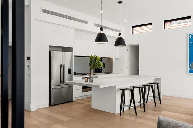 Bright contemporary open-plan kitchen with white full-height cabinetry, a large white island with integrated seating, stainless steel refrigerator, pale timber flooring, and two black pendant lights. A hallway or adjoining space is visible beyond the kitchen.