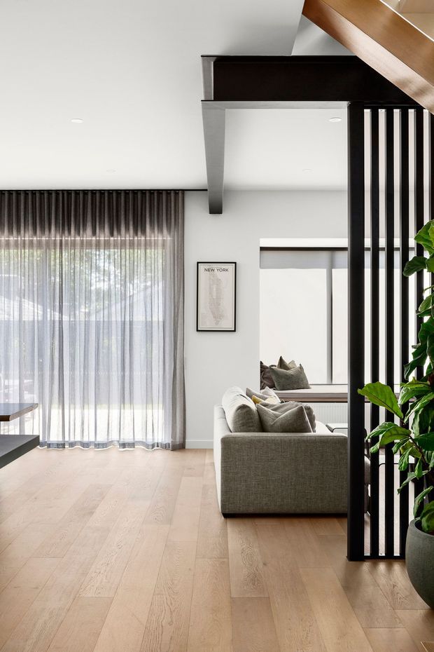 A bright contemporary interior showing an open-plan living room with a large grey sofa, pale timber flooring, sheer full-height curtains, white walls and ceiling, and a black vertical slatted partition. A timber stair or upper-level structure crosses the ceiling, while a large indoor plant adds greenery at the right. The space has abundant natural light and a calm, refined, neutral palette.