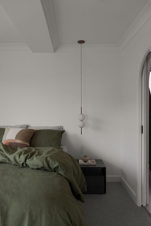 A calm, contemporary bedroom with a low bed dressed in muted olive-green bedding and layered neutral pillows. The room has pale grey-white walls, a grey carpet, white decorative ceiling mouldings, and a black bedside table with books and a small decorative object. A sculptural double-globe pendant hangs beside the bed, while an arched glazed opening or doorway is visible at right.