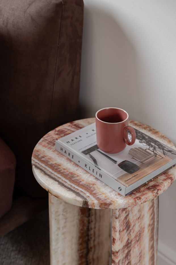 Close-up interior vignette of a small round stone side table with pronounced reddish-brown and cream veining, positioned beside a deep brown upholstered chair. A muted terracotta mug and design book sit on the tabletop against a pale neutral wall.