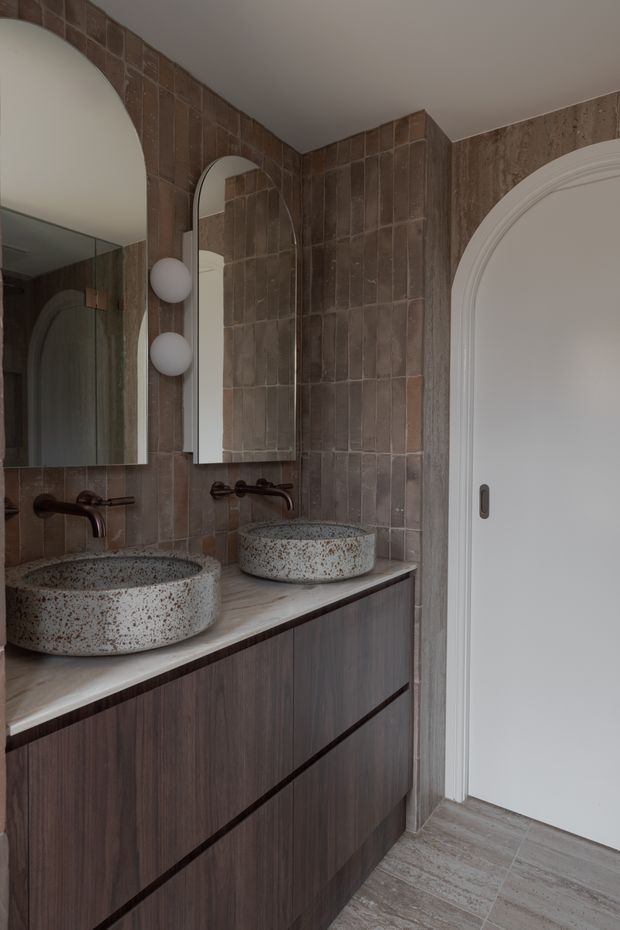 A refined bathroom vanity featuring two round speckled stone vessel basins on a pale stone benchtop, dark timber cabinetry, aged bronze wall-mounted taps, and a textured brown tile wall. Arched mirrors, spherical wall lights, and a large arched white door add sculptural architectural detail. The palette is warm, earthy, and subdued.