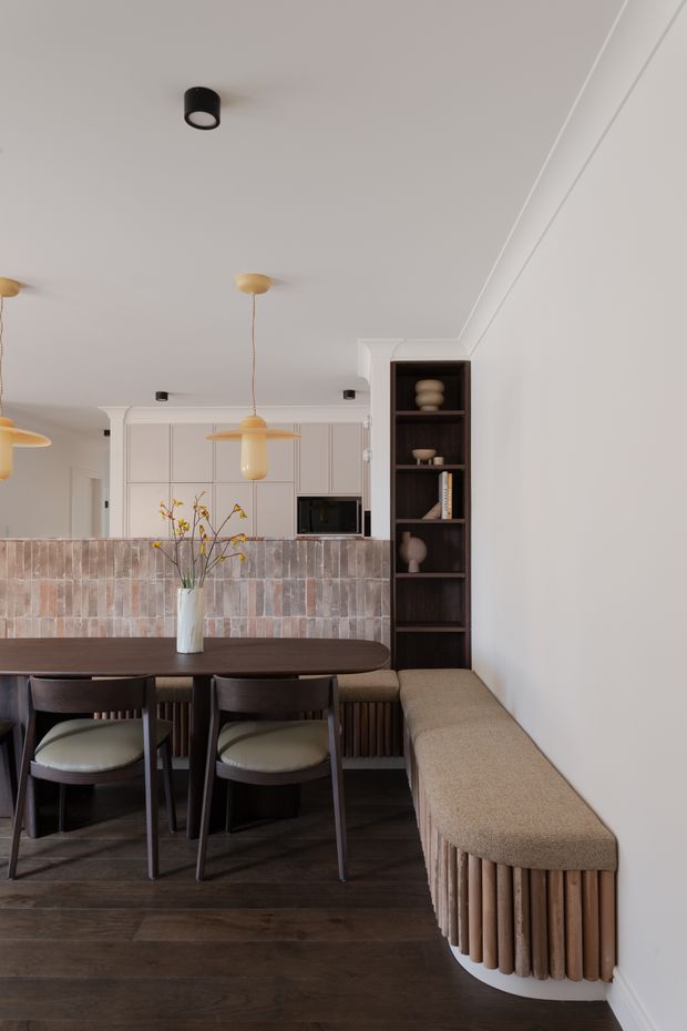 A warm, refined open-plan dining area with a dark oval dining table, upholstered dining chairs, and a built-in curved banquette with vertical timber slat detailing. Behind the dining space is a compact kitchen with pale cabinetry, a dark microwave, a textured muted pink and grey tiled splashback, and dark open shelving displaying ceramics. Pendant lights, dark timber flooring, and soft neutral finishes create a contemporary, earthy interior.