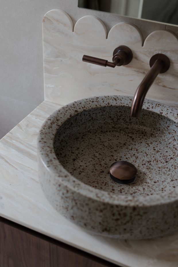 Close-up of a refined bathroom vanity featuring a large round handcrafted vessel basin in speckled brown and cream ceramic, set on a pale veined stone countertop with matching curved scalloped backsplash. A dark bronze wall-mounted faucet and separate control are visible, with a small dark soap dish inside the basin. The palette is warm, earthy, and tactile.
