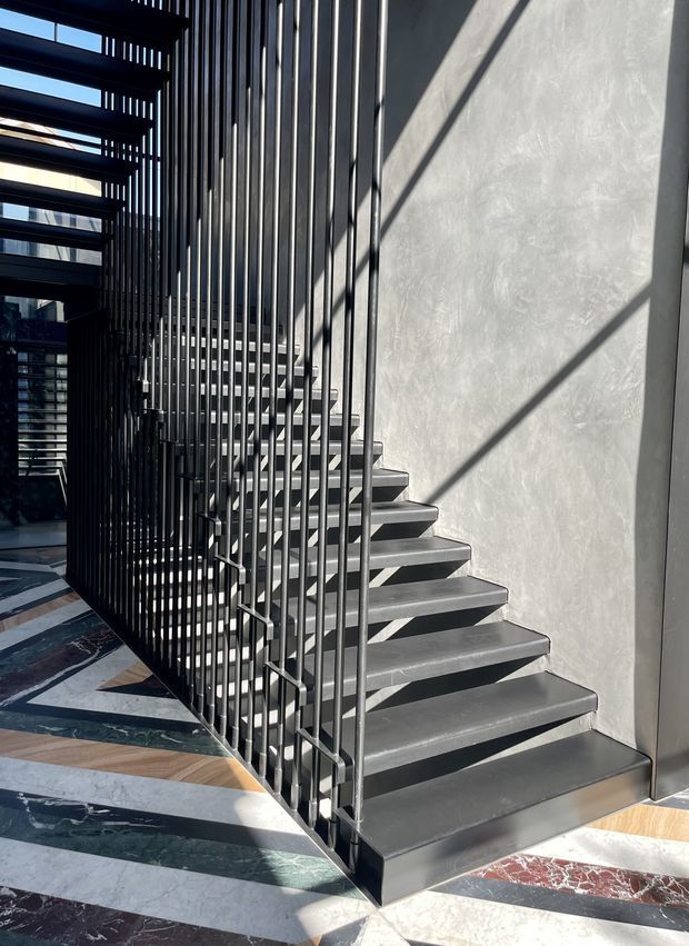 A contemporary interior stairway with dark metal floating treads and slender vertical balustrade rods. The staircase rises beside a tall smooth grey plaster wall, with strong sunlight casting geometric shadows across the wall and steps. The floor below features a dramatic multi-material geometric pattern incorporating white, grey, green, brown and red stone-like surfaces.