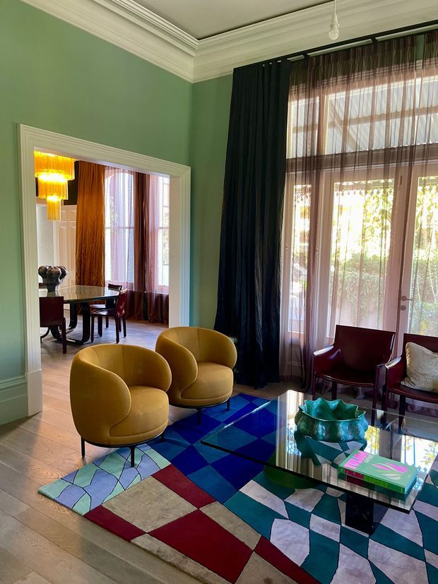 A richly styled living room in a heritage-style apartment or house, featuring sage green walls, ornate white ceiling cornices, tall glazed French doors/windows with layered sheer and dark curtains, mustard-yellow curved lounge chairs, burgundy armchairs, a reflective glass coffee table, and a bold geometric multicolour rug. A connected dining room is visible through the wide doorway, with a dark dining table, chairs, and a statement pendant light.