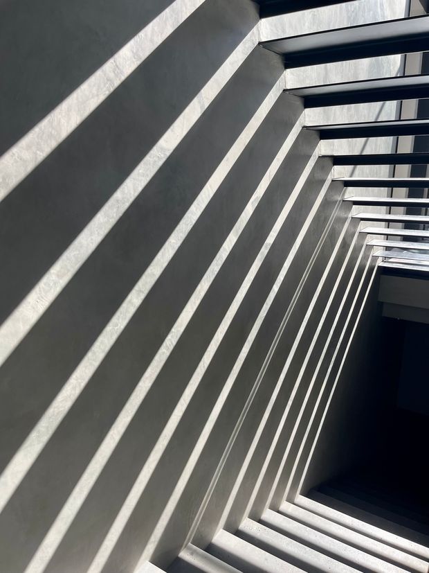 Abstract architectural view looking along a stairwell or vertical circulation space, with repeated dark metal beams and concrete or plastered walls creating strong diagonal shadows and a dramatic geometric rhythm. Bright natural light enters through the overhead openings, contrasting with the deep shadowed void below.