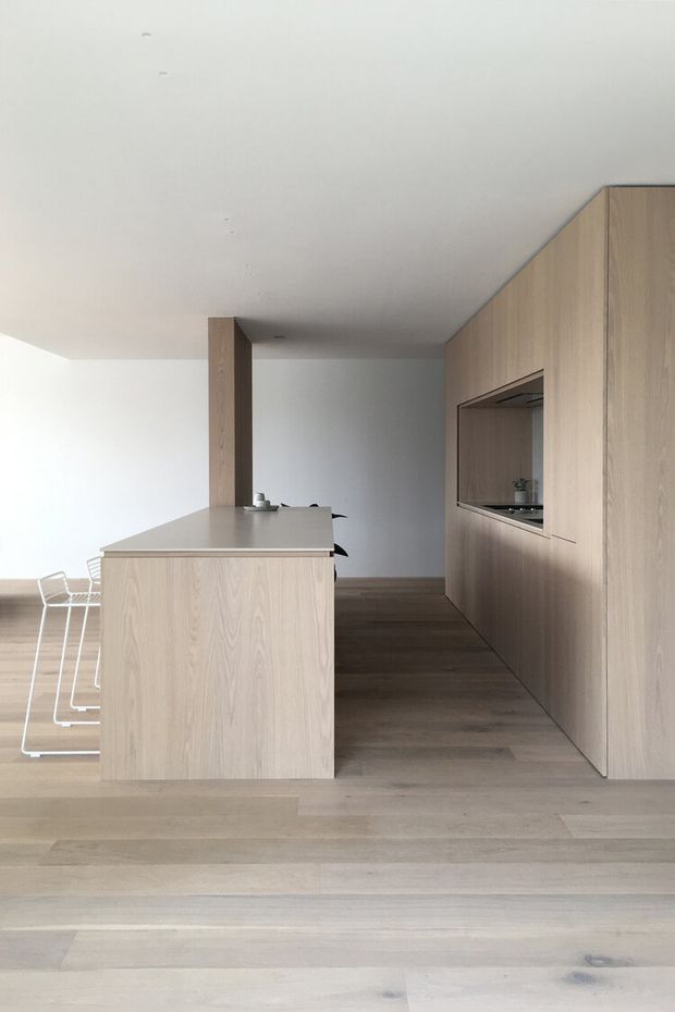A minimalist kitchen with pale timber cabinetry, a central island with a smooth light benchtop, integrated appliances, and dark timber flooring. White dining chairs are partially visible at the left, while a long wall of full-height timber joinery creates a clean, streamlined composition. The room is softly lit with a restrained neutral palette and minimal decoration.