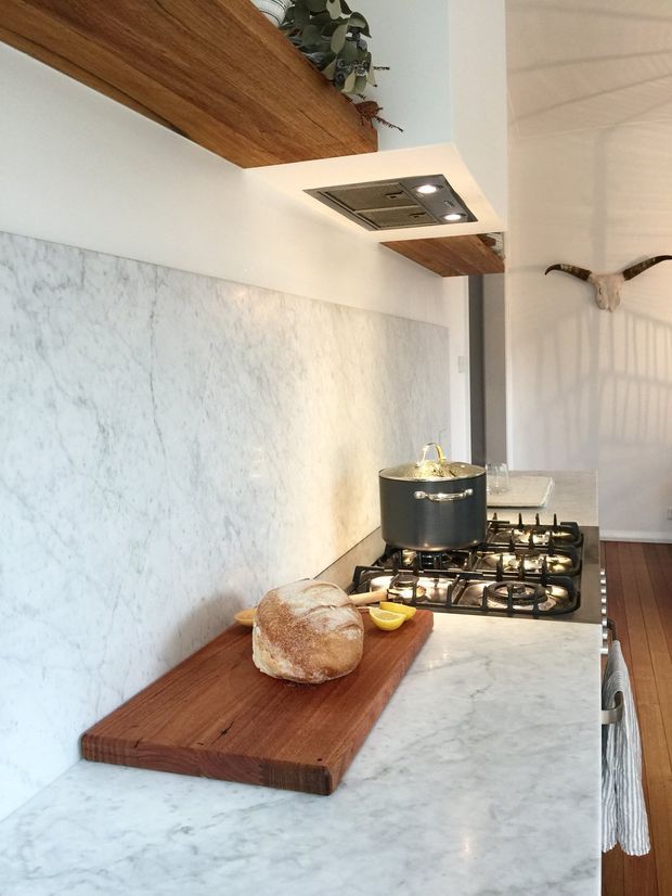 A bright, narrow contemporary kitchen featuring a white marble-look splashback and benchtop, dark timber overhead shelving, and a stainless-steel gas cooktop with integrated rangehood. A timber chopping board with bread sits on the island or work surface, while a decorative cattle skull and pendant greenery add rustic character. Timber flooring and a clean white palette create a warm, refined feel.