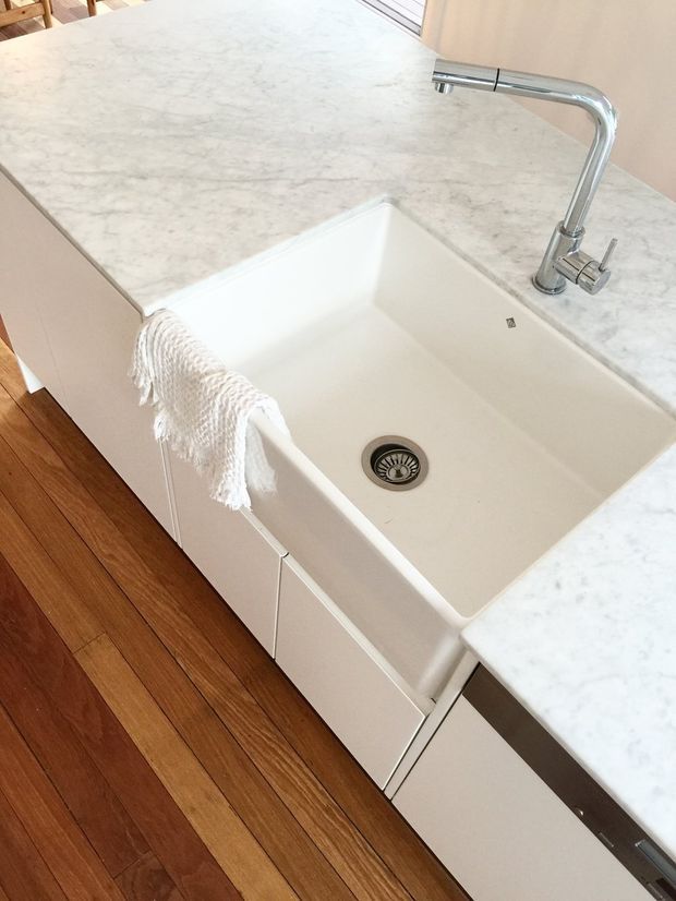 Close-up view of a minimalist kitchen sink area with a large white farmhouse-style ceramic sink, chrome gooseneck tap, pale veined stone benchtops, white cabinetry, and warm timber flooring.