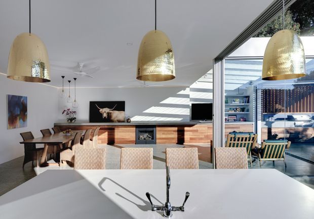 Bright open-plan kitchen and dining area with a large white island in the foreground, integrated sink and chrome tapware, timber cabinetry along the rear wall, and a built-in barbecue or fireplace feature. A dining table, upholstered chairs, pendant lights, and artwork sit to the left, while full-height glazing opens toward an outdoor entertaining area with built-in shelving and seating. The space has a clean contemporary design with warm timber accents and abundant natural light.