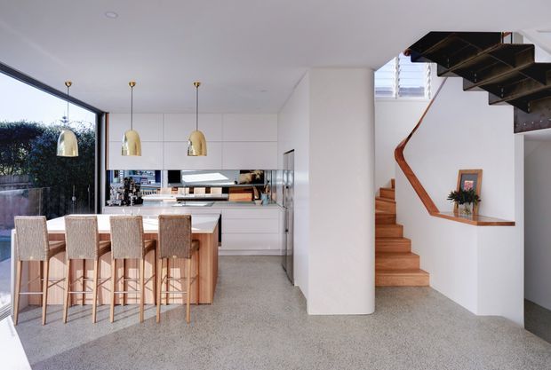 Bright open-plan kitchen and stair hall with a long island breakfast bar, four woven bar stools, white flat-panel cabinetry, mirrored splashback, and brass pendant lights. A timber stair rises beside the kitchen with a sculptural timber handrail and integrated display shelf. Large glazing opens the room toward greenery, while the floor and walls are kept light and neutral.