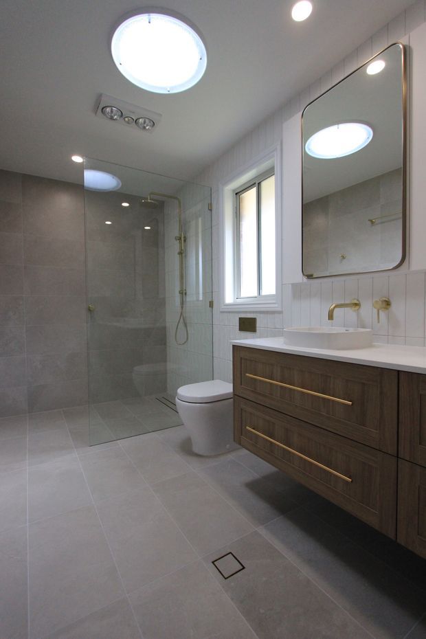 A contemporary bathroom with a walk-in glass shower, wall-hung toilet, floating dark timber vanity, white vessel basin, and brushed brass tapware and hardware. Large-format grey floor and wall tiles, white vertical wall tiles, a rounded-corner mirror, skylight, and bright recessed lighting create a clean, refined fit-out.