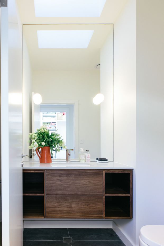 Bright contemporary bathroom vanity with a wall-to-wall mirror, white solid-surface benchtop, dark timber-grain floating cabinetry, open shelving, black floor tiles, skylight, and twin wall-mounted globe lights. A toilet is partially visible at the lower right, and a window is reflected in the mirror.
