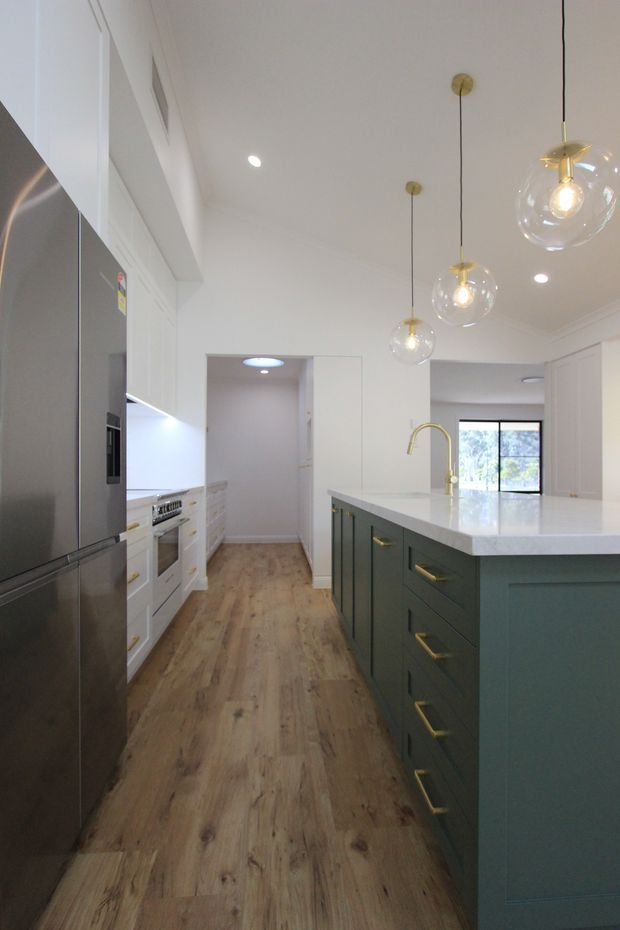 Bright contemporary kitchen viewed along a central aisle, featuring white perimeter cabinetry, a large dark green island with a white stone benchtop, integrated stainless-steel refrigerator, timber-look flooring, brass hardware and tapware, and three clear glass globe pendant lights. A dining or living area is visible beyond the island through the open-plan space.