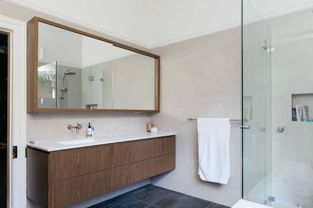 A bright contemporary bathroom featuring a wall-mounted dark timber vanity with a white integrated basin, a large timber-framed mirrored cabinet, pale large-format wall tiles, dark floor tiles, and a frameless glass walk-in shower with recessed shelving. Chrome tapware and hardware provide subtle contrast.