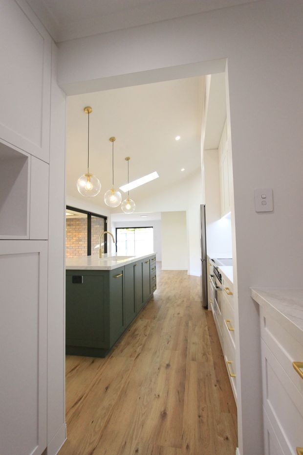 A bright, narrow contemporary kitchen viewed through a wide doorway. The room features a dark green island with a white stone benchtop, white built-in cabinetry, warm timber-look flooring, black-framed glazing, exposed brick at the rear, and three globe pendant lights over the island.