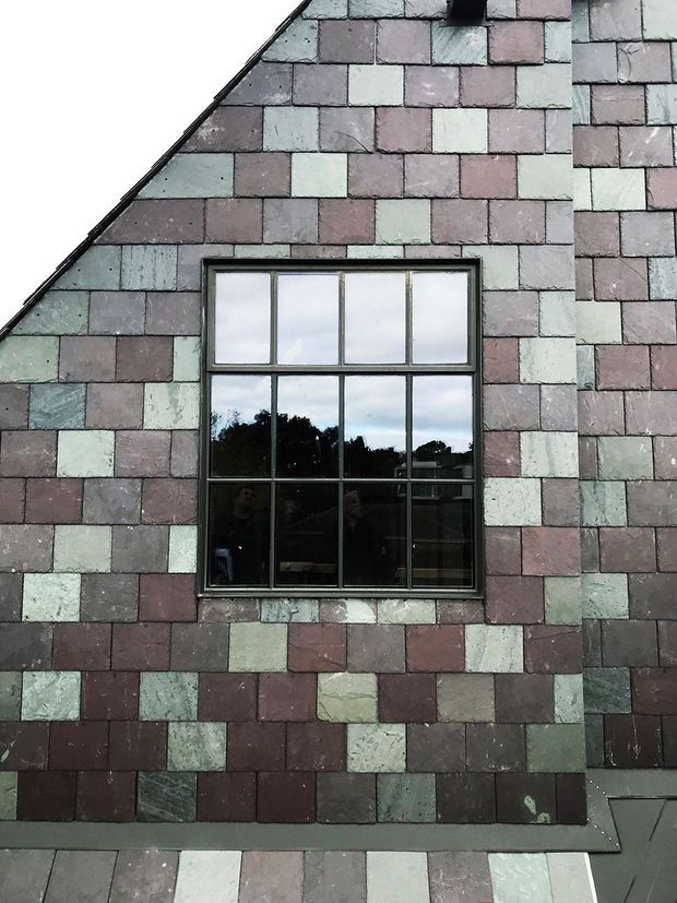 Close-up exterior view of a steeply gabled building facade clad in small, irregular rectangular slate tiles in muted pink, mauve, grey, and green tones. A dark metal-framed multi-pane window is centered in the facade, reflecting trees and sky. The image focuses on the roof-like wall cladding and window detail.