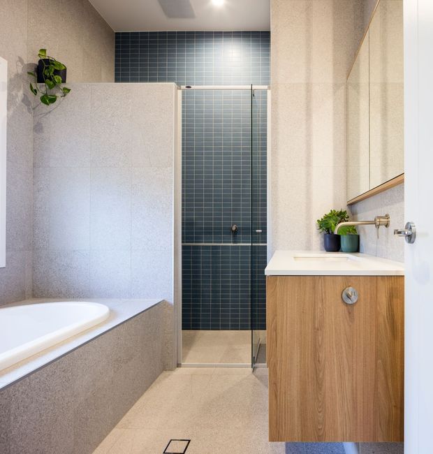 Contemporary bathroom with a freestanding-style bathtub partially visible at left, a walk-in glass shower with dark blue grid-pattern wall tiles, and a timber vanity with a white integrated basin. Large-format light beige stone-look tiles cover the walls and floor, with a frameless mirror and small indoor plants adding warmth.