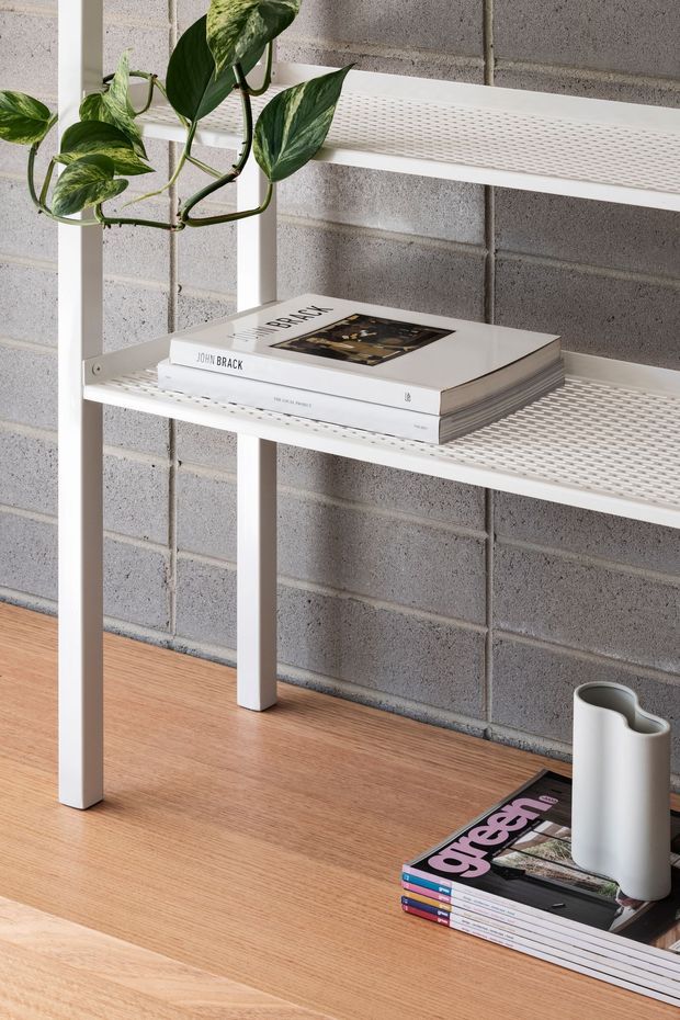 Close-up interior detail of a minimalist white open shelving unit with perforated metal shelves, positioned against a grey painted concrete block wall. A trailing variegated plant, stacked design books and magazines, and a sculptural white vase sit on and around the shelving. The unit stands on a warm timber floor, creating a clean contemporary setting with natural greenery and restrained styling.