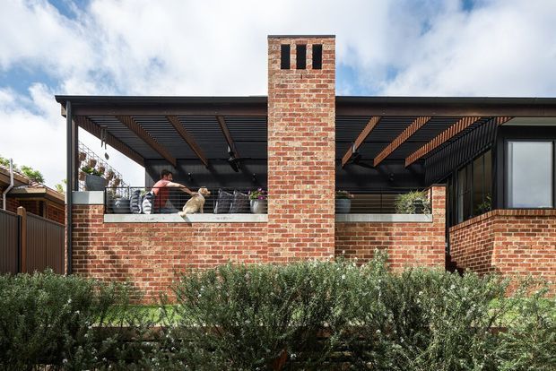 Exterior view of a contemporary brick residence with a covered open-air entertaining area, exposed timber pergola detailing, dark metal framing, large glazing, and a prominent brick chimney. Landscaping partially screens the lower facade.