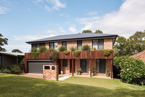 Exterior view of a contemporary two-storey brick house with a low-pitched metal roof, dark-framed windows, integrated garage, and a timber-clad upper-level balcony planter. The landscaped front yard and neighbouring residential buildings are visible under a bright sky.