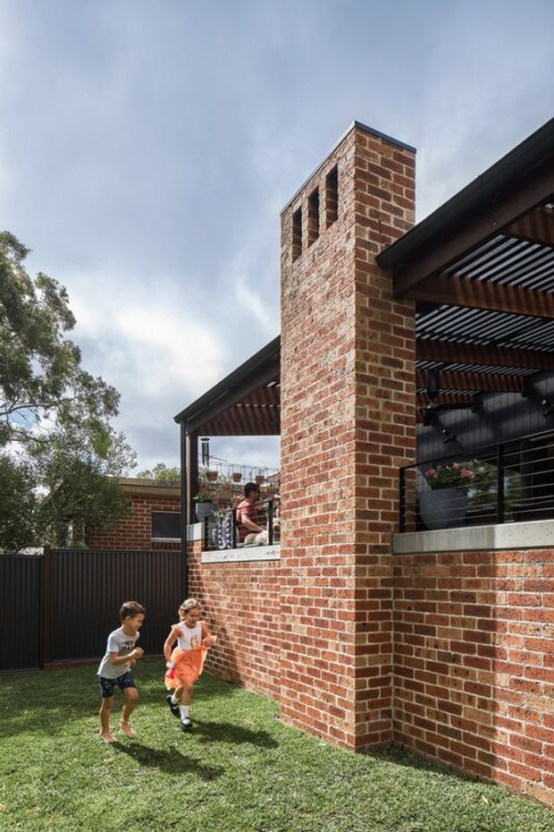 Exterior view of a contemporary residential extension with prominent red brickwork, a tall brick chimney or masonry feature, dark metal-framed openings, and a covered outdoor terrace with slatted pergola roofing. The building sits beside a grass-covered yard and established trees.