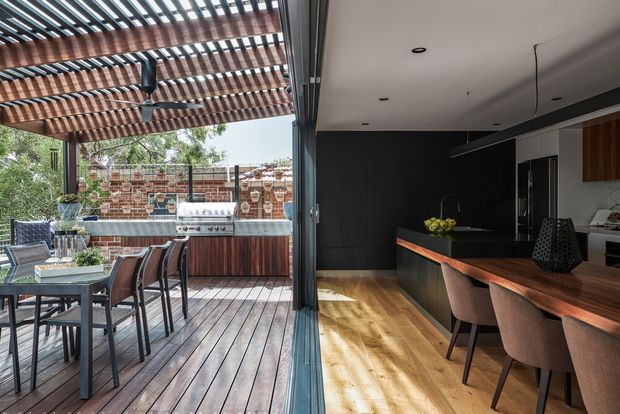 A contemporary indoor-outdoor entertaining space viewed through fully opened sliding glass doors. The left side shows a timber deck with dining table, chairs, barbecue, and a covered pergola with exposed timber beams and slatted roof. The right side is a dark, refined kitchen with a long island, warm timber flooring, dark cabinetry or wall panelling, timber benchtop detailing, pendant lighting, and bar stools. Brick screening and garden vegetation are visible beyond the deck.