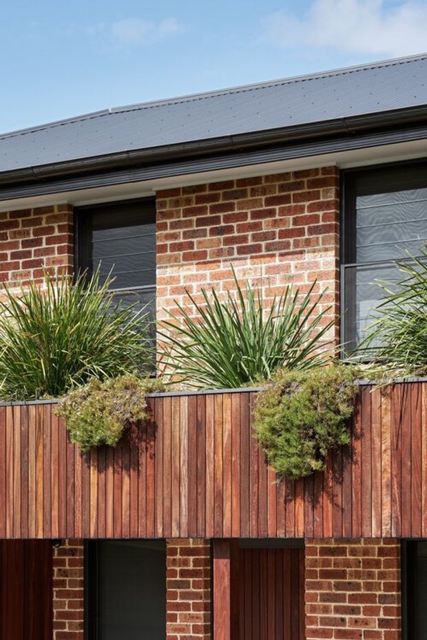 Close-up exterior view of a multi-level brick residential facade with dark-framed windows and doors beneath a grey metal pitched roof. A projecting vertical timber planter or balcony screen spans the middle level, containing grasses and shrubs. The composition highlights the brickwork, timber cladding, metal roofing, and integrated planting in bright daylight.