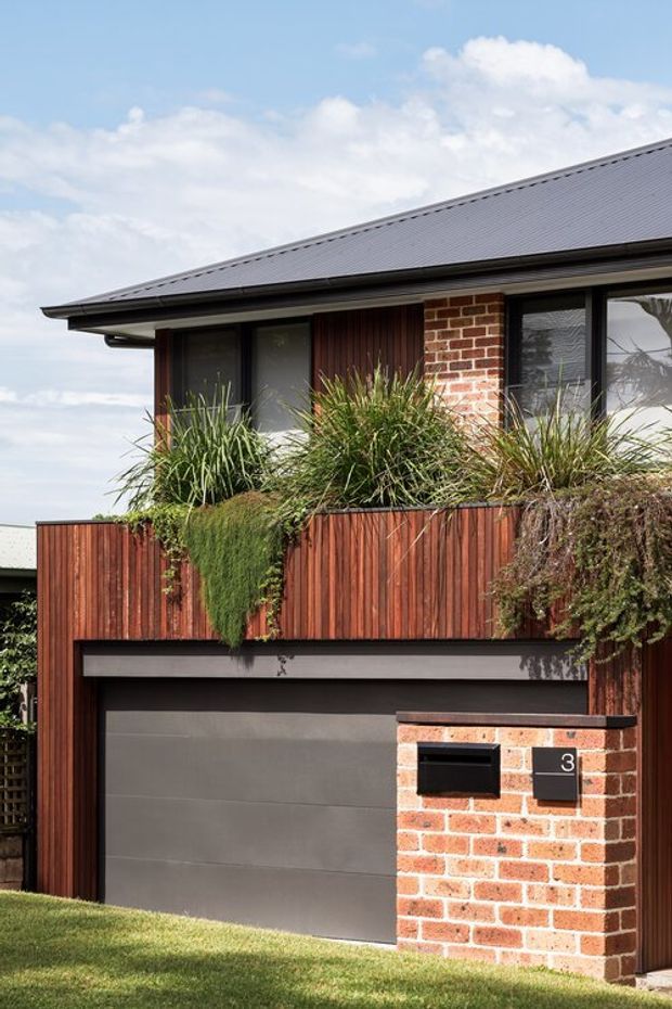 Exterior view of a contemporary residential townhouse or house facade. The image shows a dark grey metal pitched roof with broad eaves, red brick walls, black-framed windows, and a dark grey sectional garage door. Vertical reddish timber screening forms a balcony or privacy fence above the garage, with lush ornamental planting spilling over it. A brick mailbox pillar and the number 3 are visible at the front boundary.