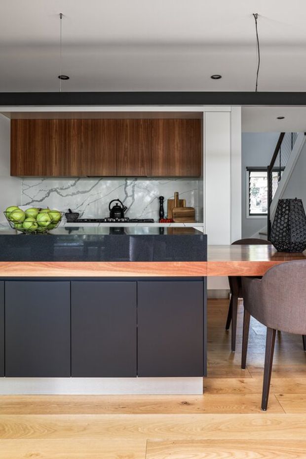 A contemporary open-plan kitchen viewed from the adjoining dining area. The kitchen features dark matte island cabinetry, a polished dark stone benchtop, warm walnut overhead cabinetry, and a dramatic white marble-look splashback with grey veining. A timber dining table and upholstered dining chairs occupy the foreground, with a stairway visible beyond the kitchen. The space has pale timber flooring, white walls, recessed downlights, and a bold black horizontal structural or ceiling element.