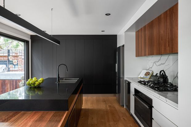 Contemporary galley-style kitchen with dark matte cabinetry, a large black stone island with integrated sink and gooseneck tap, warm timber flooring and island detailing, white cabinetry, a veined marble-look splashback, stainless-steel appliances, and a timber overhead cabinet. The space has strong natural light from a large window and a clean, refined material contrast.