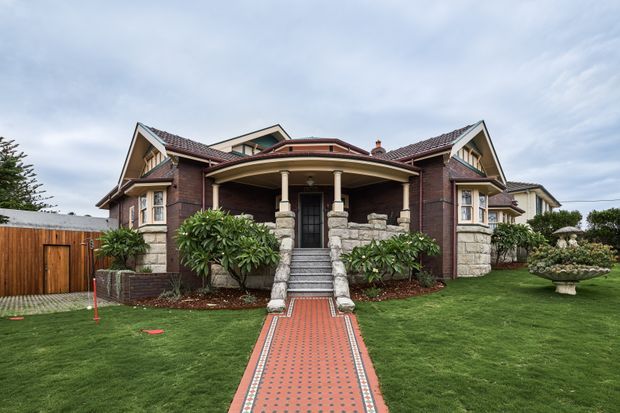 Front exterior view of a large traditional brick residence with prominent gabled roof forms, a curved covered entry porch supported by columns, stone-clad porch piers and stair walls, and a patterned red tiled pathway leading through a manicured front lawn. Timber fencing and mature planting frame the property.