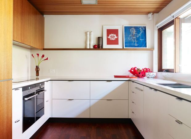 A bright, compact contemporary kitchen with white L-shaped cabinetry, integrated black appliances, a dark timber floor, and a warm timber slatted ceiling. Timber-framed glazing along the right side brings in abundant daylight. Minimal benchtops and a small collection of colourful artwork and decor create a clean mid-century-influenced interior.
