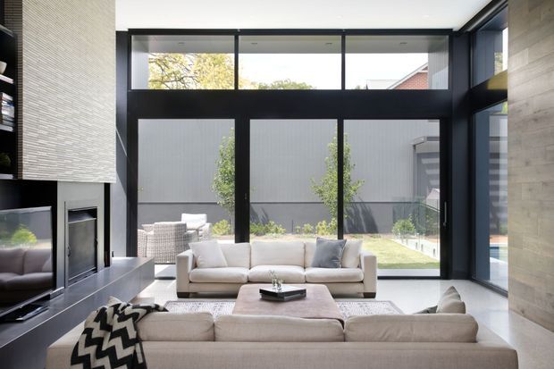 Contemporary living room with two cream sofas arranged around a low rectangular coffee table, a large black-framed glazed wall, and a built-in fireplace and media joinery. The room opens visually onto a landscaped courtyard, with a concrete-toned feature wall and abundant natural light.
