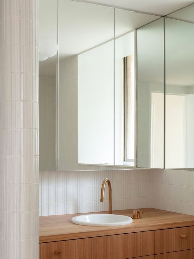 Bright, minimalist bathroom vanity with a warm timber countertop and cabinetry, white oval vessel basin, brushed brass gooseneck tap, white vertically ribbed wall tiles, and large mirrored overhead cabinets. A tiled cylindrical column and soft natural light add architectural interest.