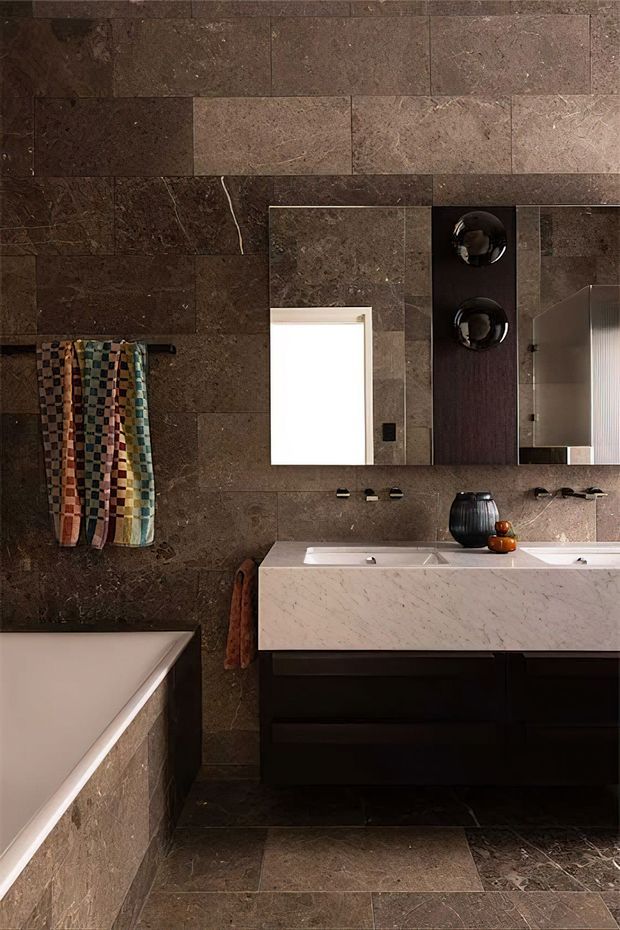 A refined contemporary bathroom featuring dark brown stone-look wall and floor tiles, a freestanding bathtub, and a floating dark timber vanity with a white marble benchtop and twin basins. A large mirror, wall-mounted fixtures, black fittings, and colourful patterned towels complete the sophisticated, moody interior.