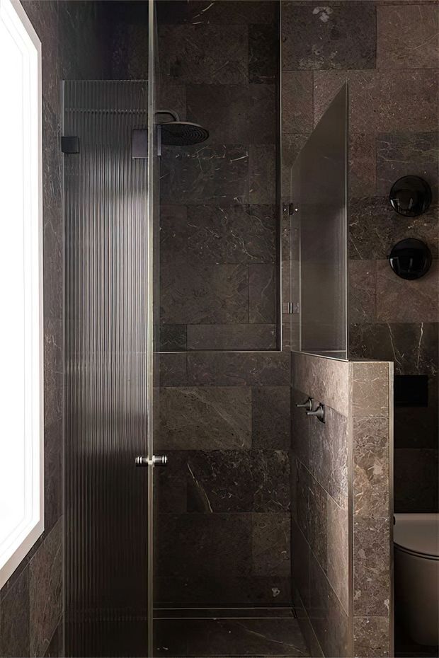 A compact, dark-toned bathroom focused on a glass-enclosed walk-in shower. Large-format brown-grey stone-look tiles cover the walls and floor, with a ribbed translucent glass shower screen, black shower fixtures, a wall-mounted control, and a narrow tiled partition. A window provides bright natural light on the left, while part of a toilet is visible at the right.