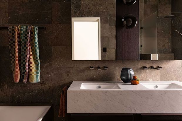 A sophisticated contemporary bathroom viewed from above, featuring a double vanity with a white marble-look benchtop and integrated basins, dark brown stone wall and floor finishes, a large frameless mirror, wall-mounted taps, and colourful hanging towels. A partial bathtub is visible in the lower-left corner.