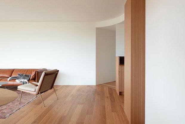 A refined open-plan living area with warm timber flooring, a tan leather sofa, a vintage-style lounge chair, and a round timber coffee table. Curved white walls and a timber-clad vertical partition lead toward a partially visible kitchen or joinery area, creating a clean, understated interior.