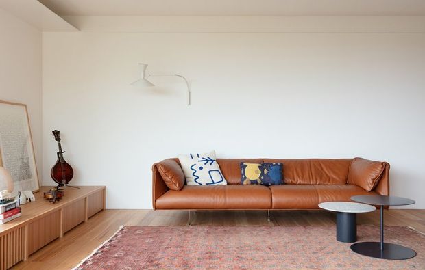 A bright, minimalist living room with a long cognac leather sofa, patterned cushions, a large vintage-style rug, and a pair of round black and white nesting coffee tables. A low timber media console lines the wall, with a guitar and framed artwork adding character. The room has white walls, timber flooring, simple wall-mounted lighting, and a calm contemporary feel.