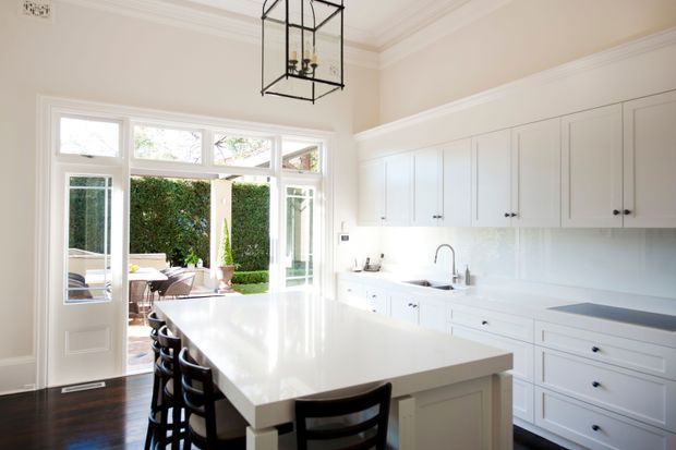 Bright, spacious traditional-style kitchen with extensive white shaker cabinetry, a large white island with seating, dark timber flooring, pale walls, and abundant glazing opening to an outdoor dining area. A black geometric pendant light provides contrast, while the overall palette is clean and refined.