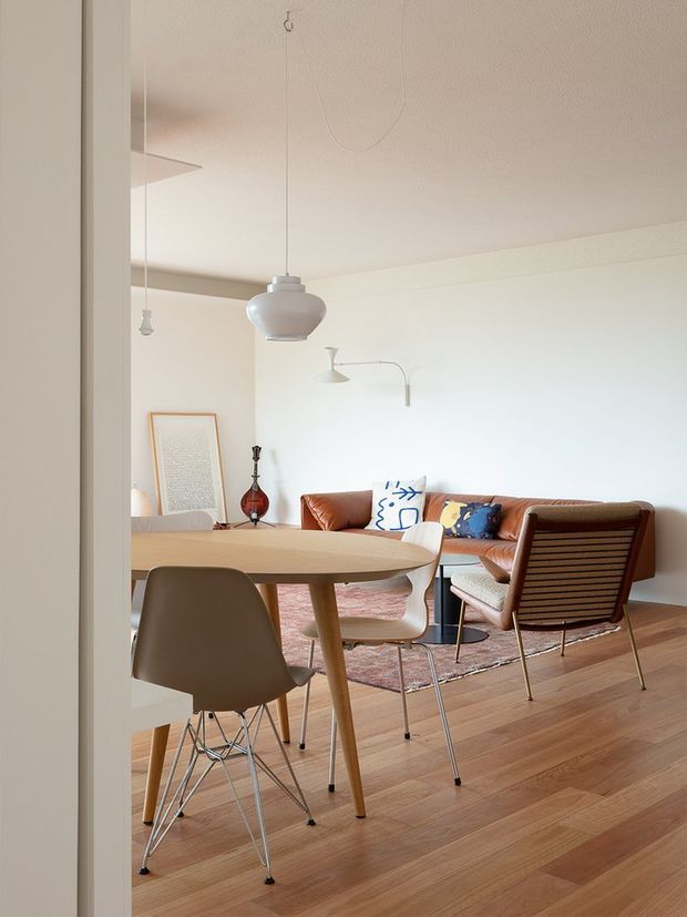 Bright, open-plan mid-century-inspired living and dining area viewed through a doorway. The room features a long light-wood dining table with molded chairs, a brown leather sofa, a vintage wood-and-leather lounge chair, a patterned rug, timber flooring, sculptural pendant lighting, and minimalist wall-mounted lighting. The palette is warm and neutral with restrained decorative accents.